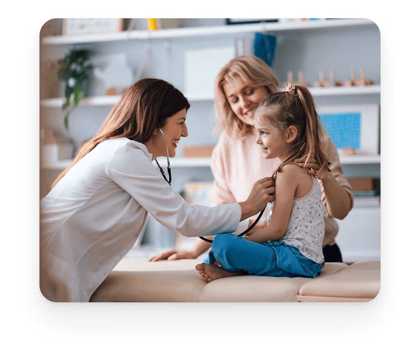 A female doctor in a white coat using a stethoscope to listen to a smiling young girl sitting on an exam table, with the girl's mother smiling behind her.