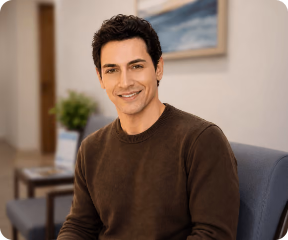 Smiling man with dark hair wearing a brown sweater sitting on a blue chair in a room with a plant and a framed picture on the wall.