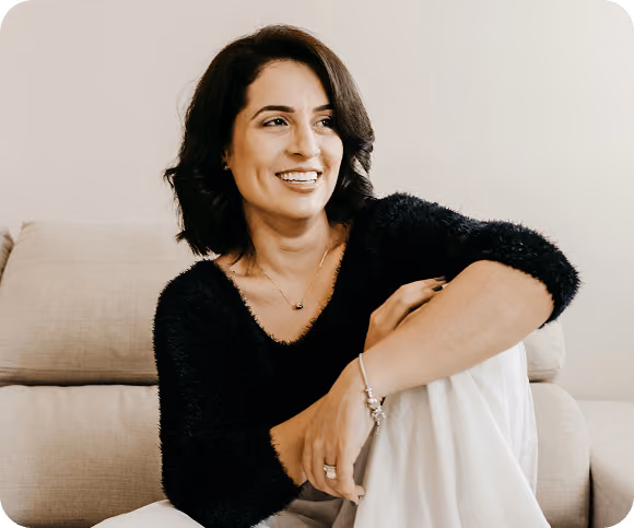 Smiling woman with dark hair wearing a black fuzzy sweater and white pants, sitting on a beige couch with one knee raised.