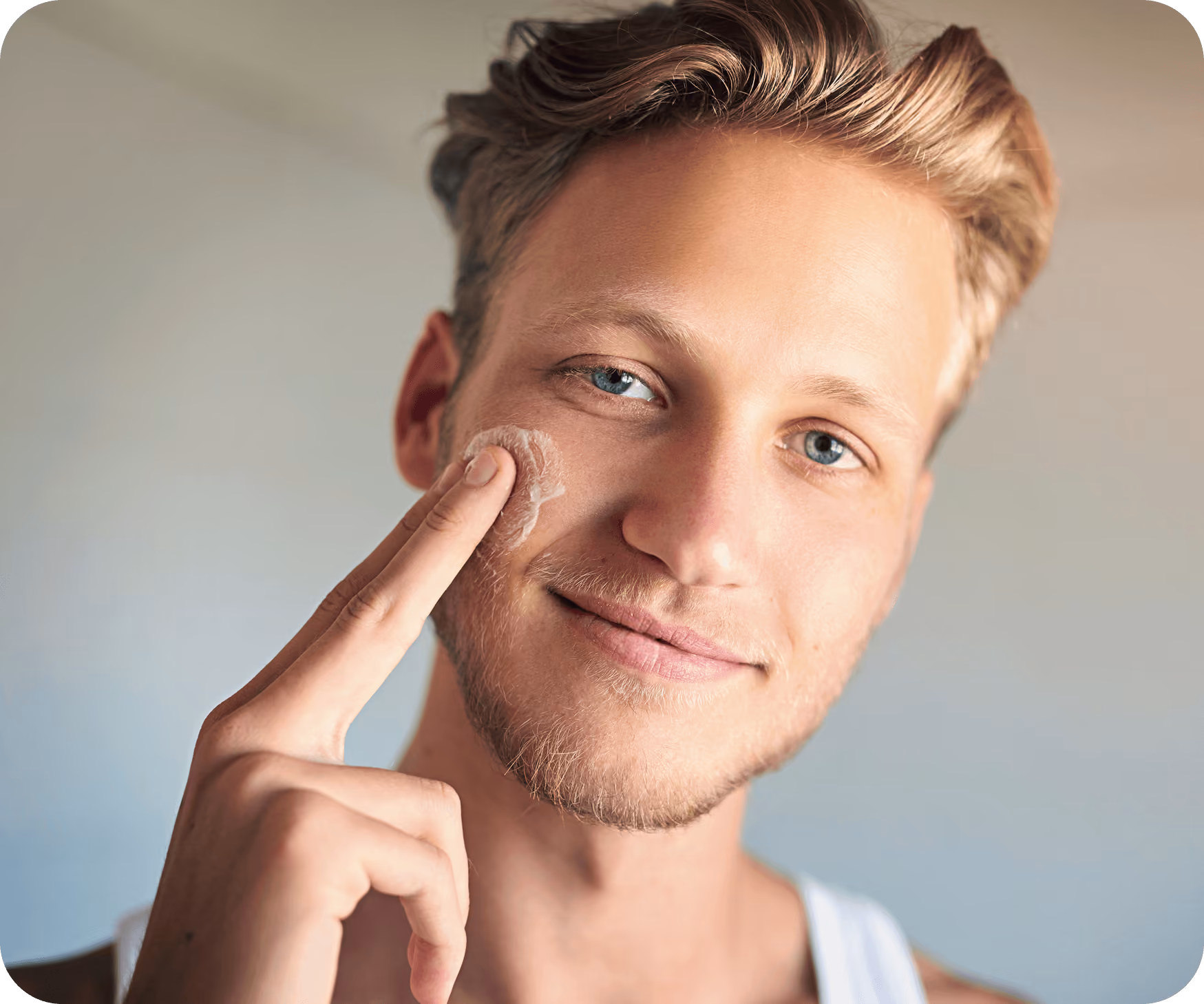 Close-up of a young man with light brown hair and blue eyes applying facial cream to his right cheek with his index finger while smiling slightly.