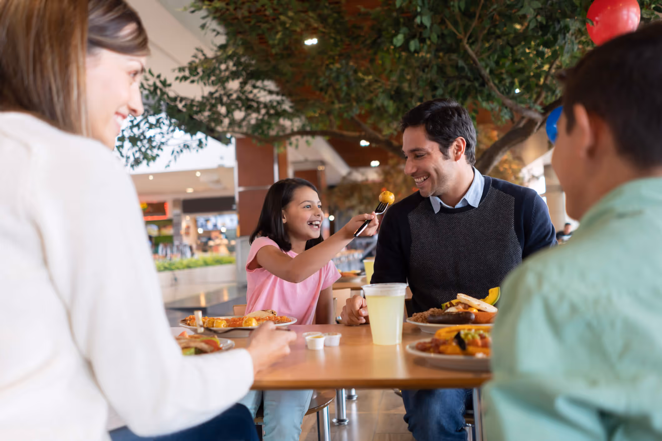 A young girl playfully feeds a man a bite of food with a fork as they smile at each other, sitting at a table with two other people and plates of food.