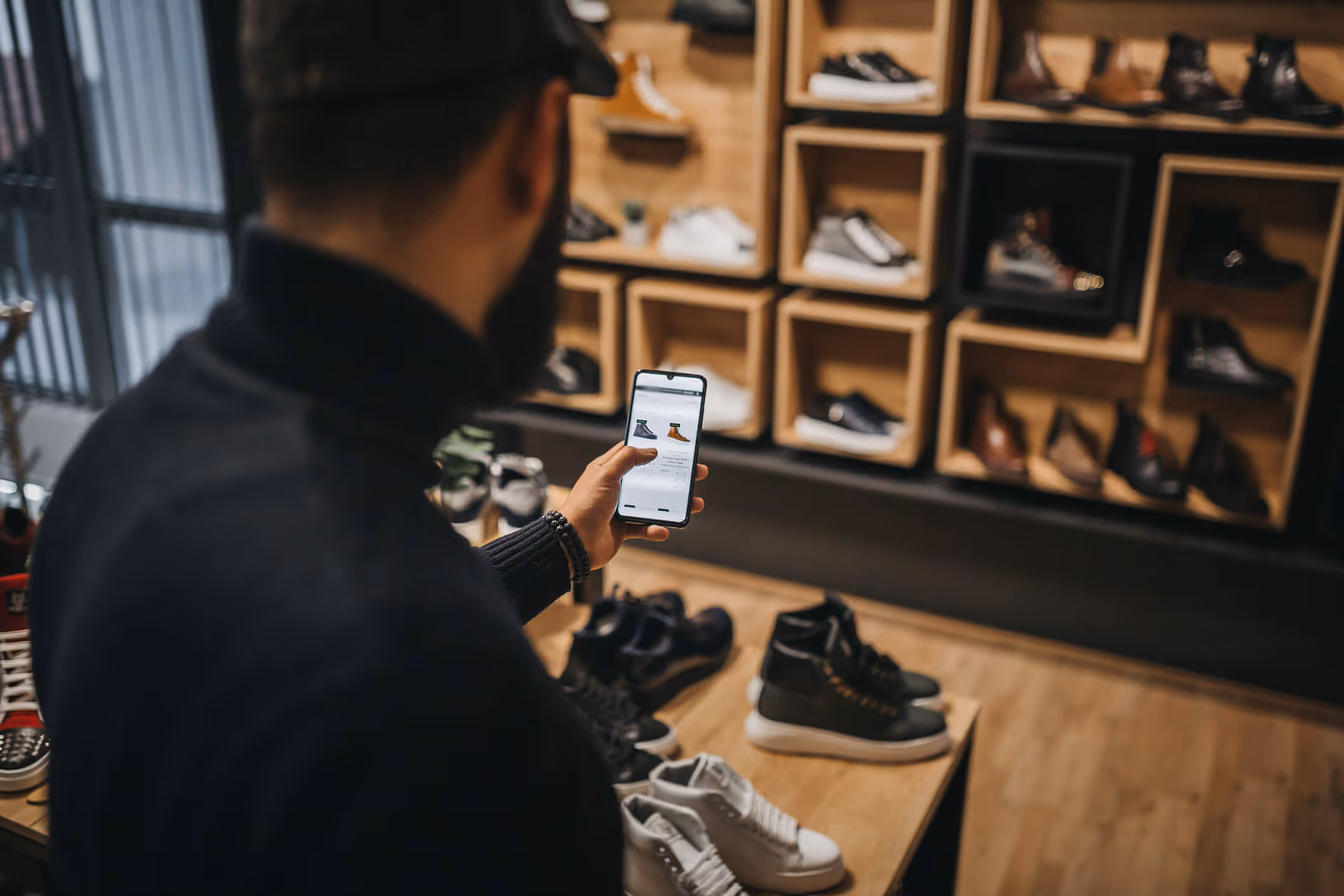 Man in a shoe store browsing boots on a smartphone with various shoes displayed on shelves and tables.