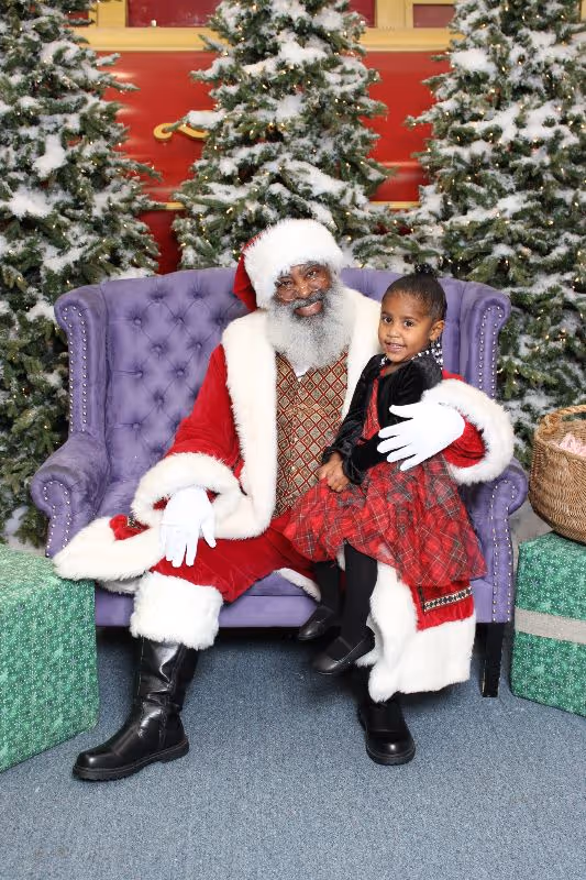 Black Santa Claus in red and white suit sitting on a purple chair holding a young girl in a red and black dress, with snow-covered Christmas trees in the background.