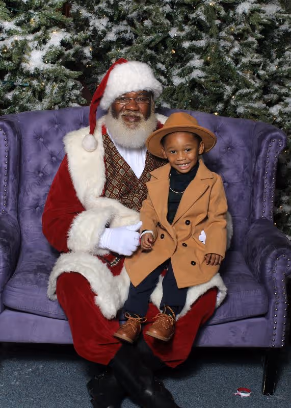 Young boy in a tan coat and hat sitting on the lap of a Black Santa Claus dressed in a red and white suit on a purple chair with snowy pine trees in the background.