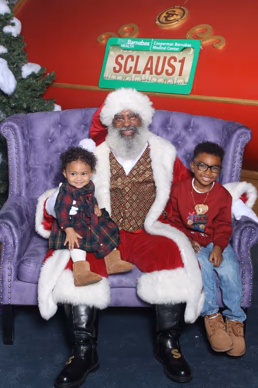 Black Santa Claus sitting on a purple velvet couch with a smiling toddler in a plaid dress on his lap and a boy in glasses and a red sweatshirt sitting beside him.