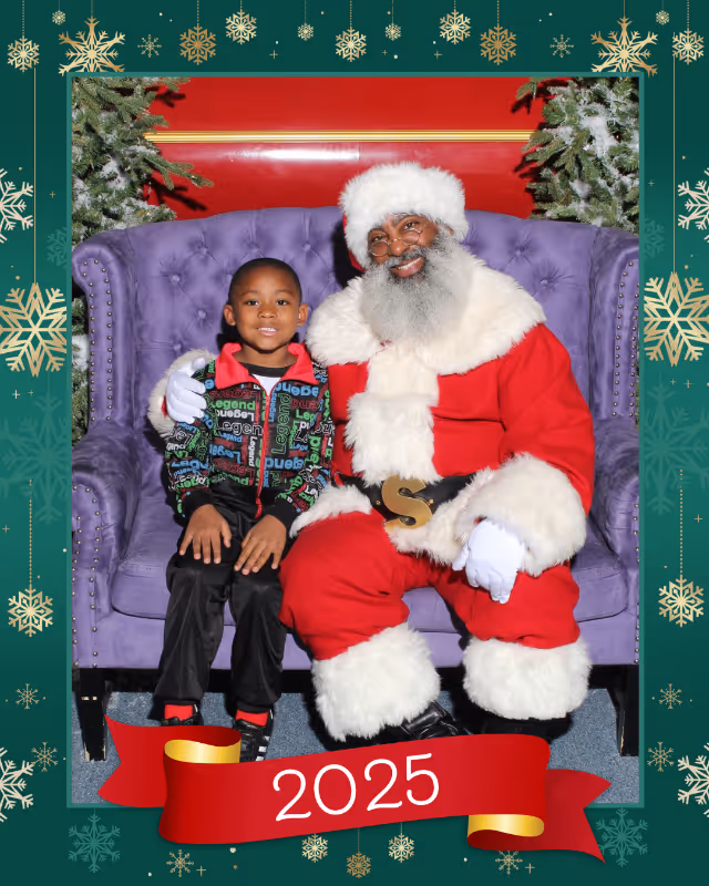 Smiling Black Santa with a gray beard sitting on a purple couch next to a young Black boy wearing a colorful jacket, with Christmas trees in the background and a red 2025 banner below.