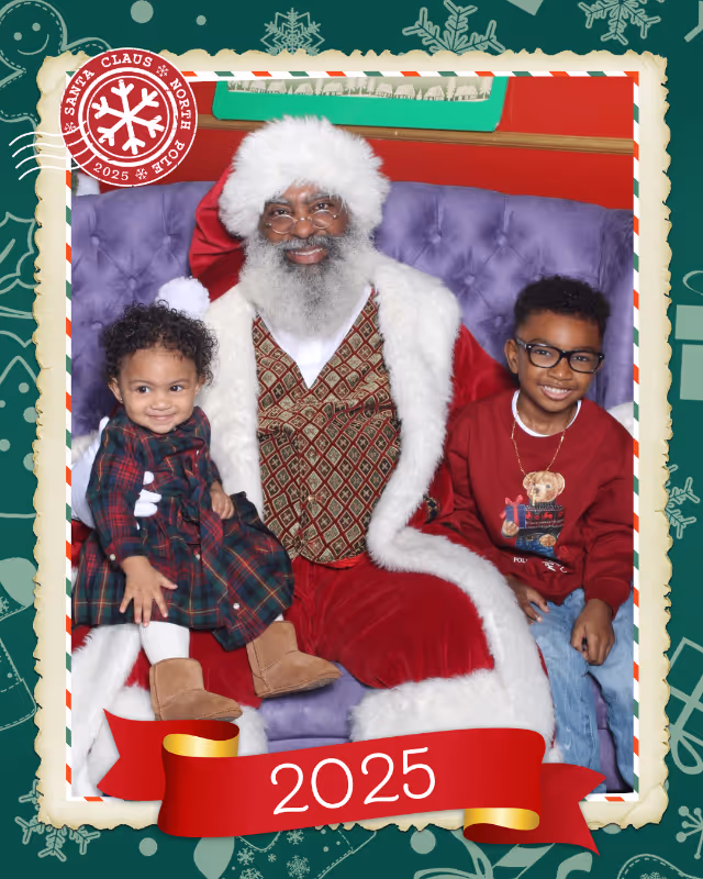 Black Santa Claus with white beard and glasses sitting on a purple velvet couch with two smiling children, a girl in a plaid dress and a boy in a red sweatshirt, with '2025' on a red ribbon below.