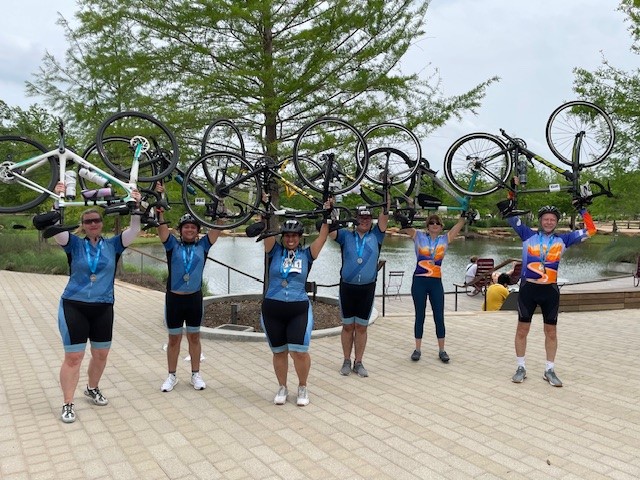 Group of six cyclists holding bikes overhead at a park near water and trees.