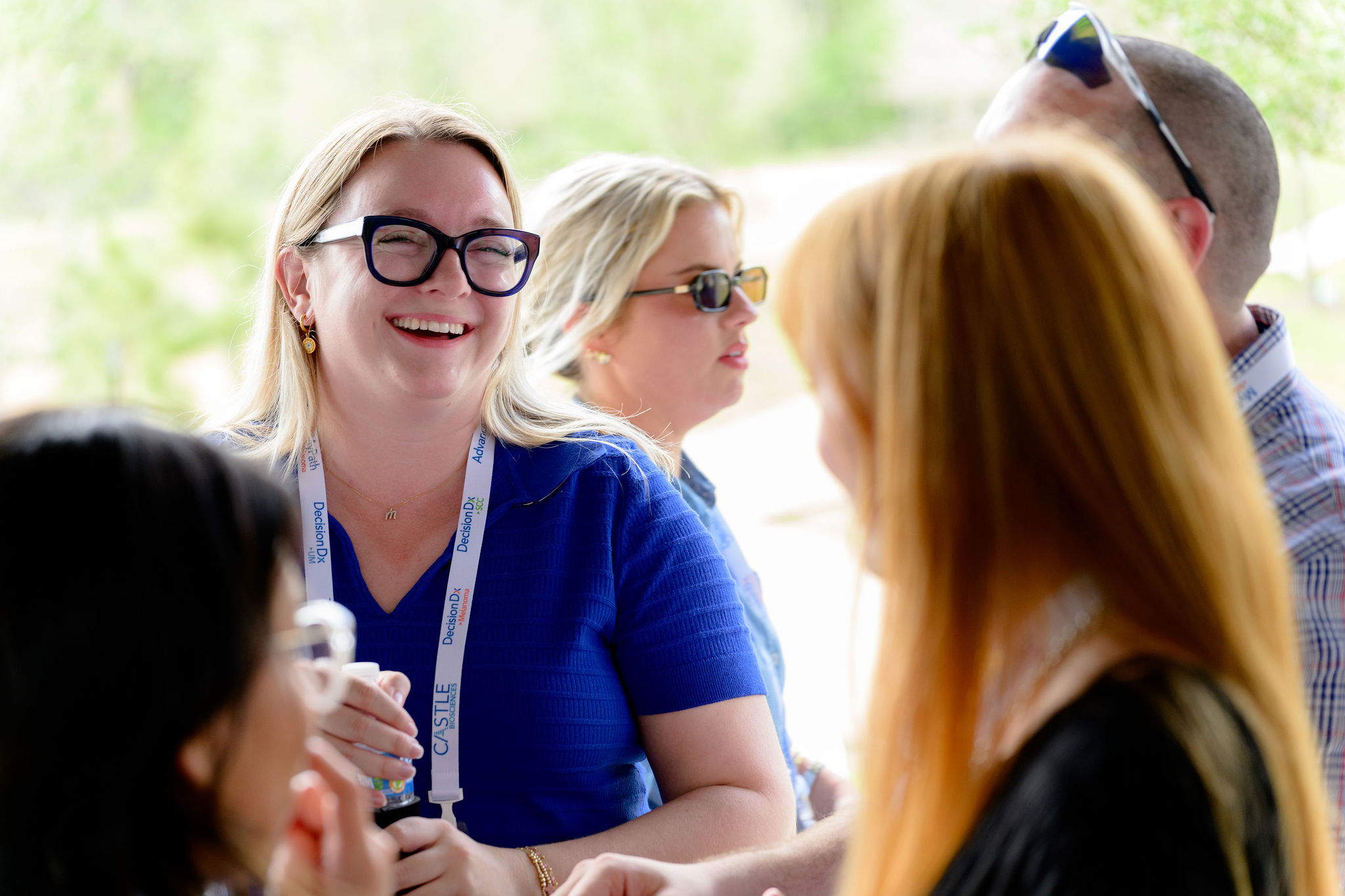 Smiling woman in blue shirt and glasses talks with three others outdoors in bright setting.