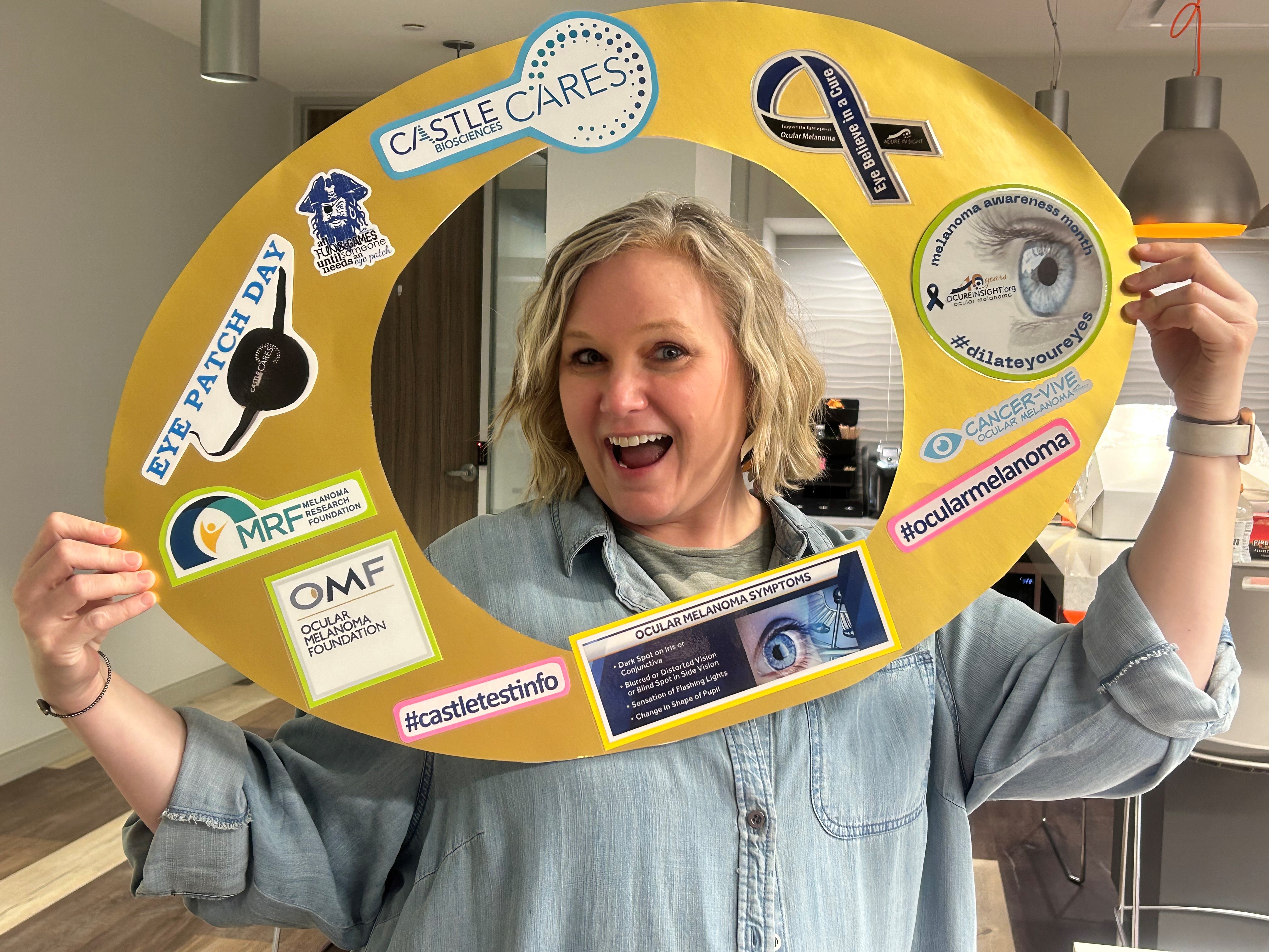 Smiling woman holding a large yellow eye patch cutout with ocular melanoma awareness stickers.