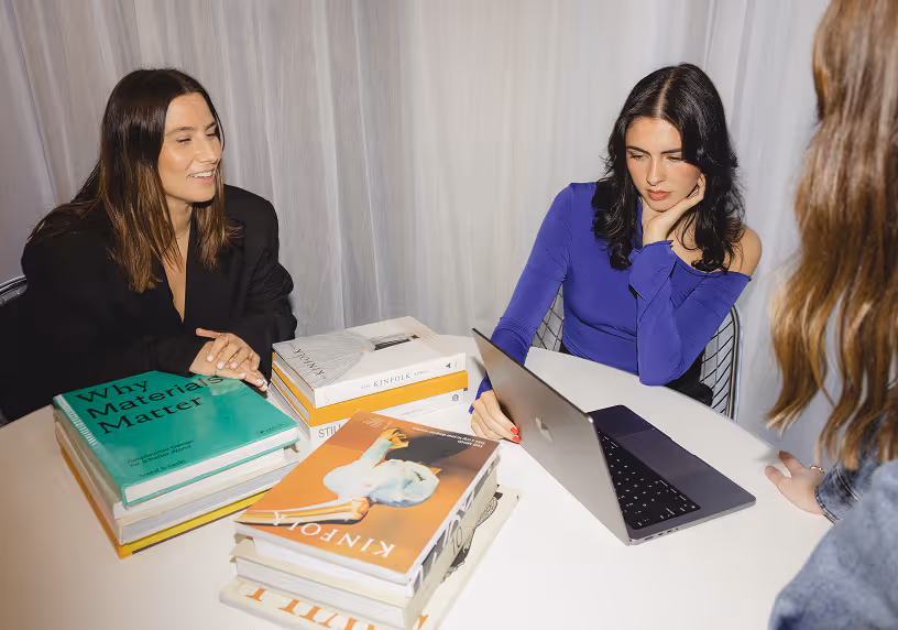 Three women at a table with books, one using a laptop as they discuss work or study.