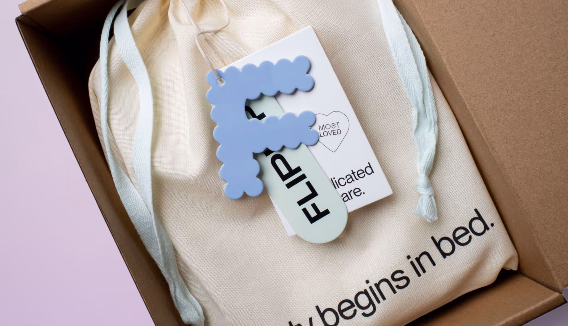 Boxed fabric drawstring bag with text 'begins in bed' and a keychain with a blue letter F.