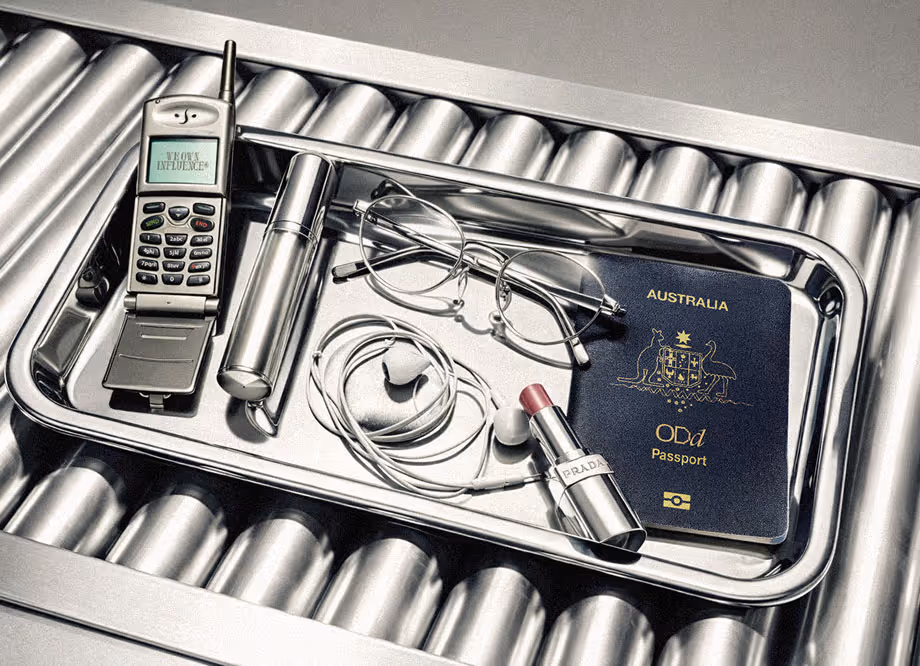Travel tray with old flip phone, lipstick, glasses, earbuds, and Australian passport on baggage scanner.