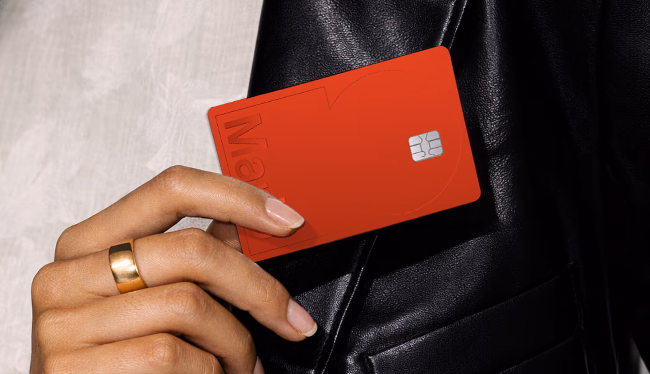 Hand with gold ring holding a red credit card against a black leather jacket.