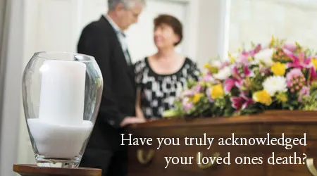 A couple stands by a casket with a candle in the foreground.
