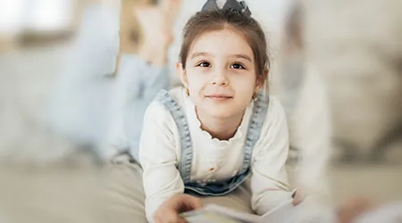 A young girl in overalls lies on a bed, smiling and reading.
