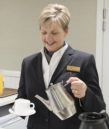 A smiling, short-haired woman in a dark suit pours coffee from a silver pot into a white cup.
