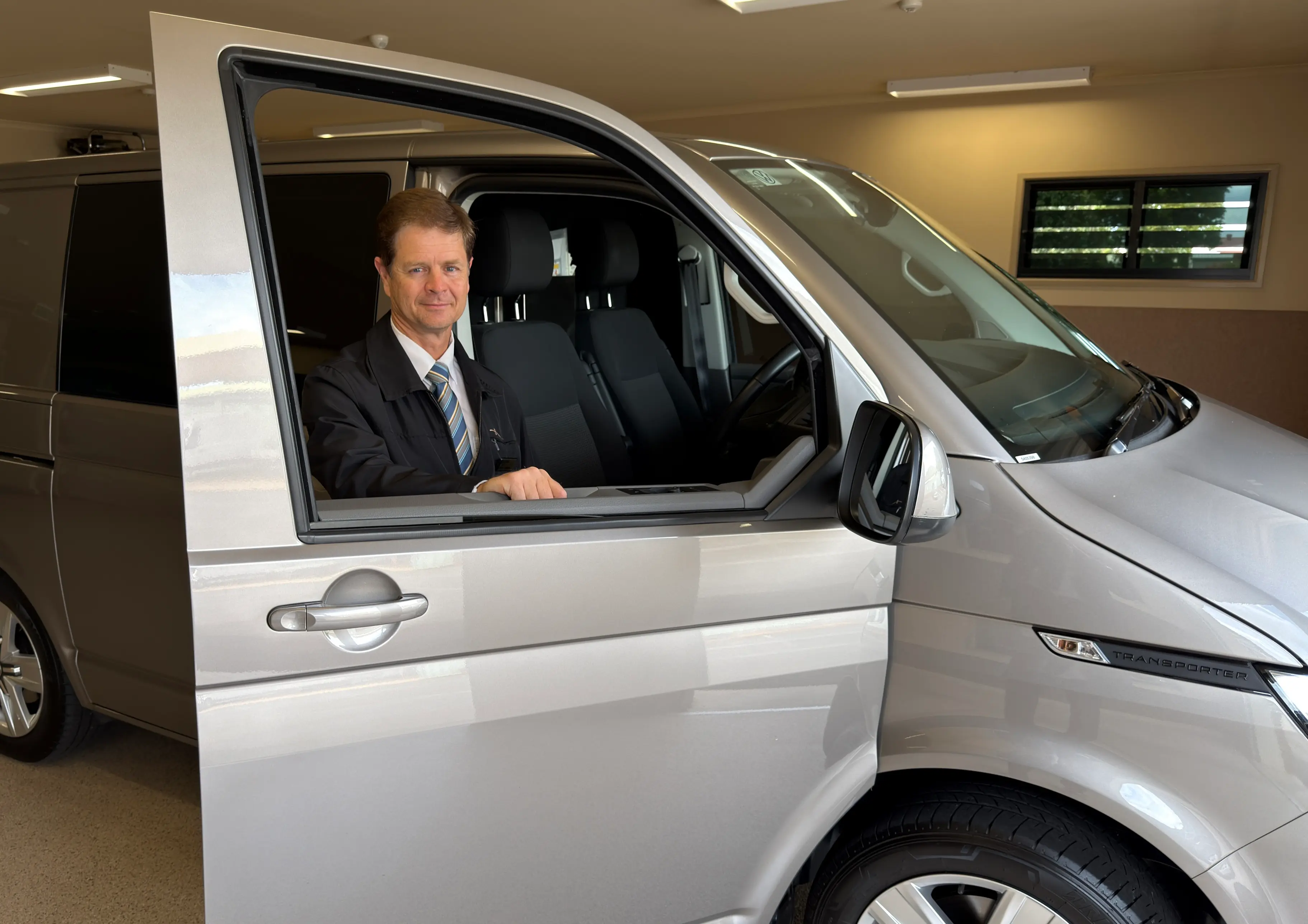 A man in a suit and tie stands in the open driver's door of a silver Volkswagen Transporter.