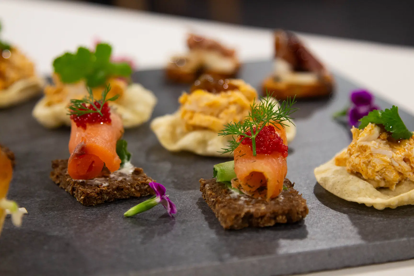 Assorted gourmet canapés with smoked salmon and toppings served on a dark slate board.