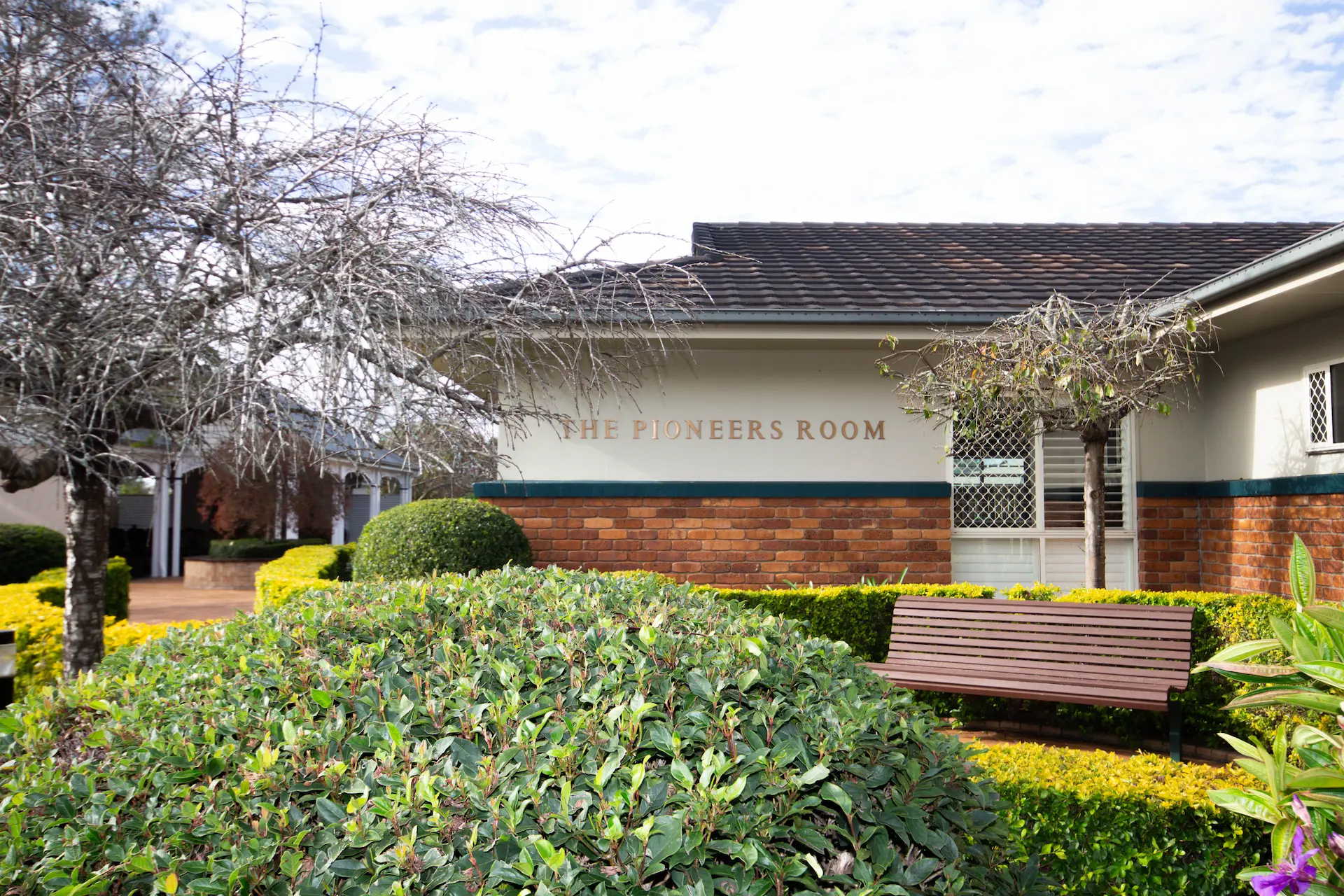 Exterior of The Pioneers Room building surrounded by green bushes and flowering trees.