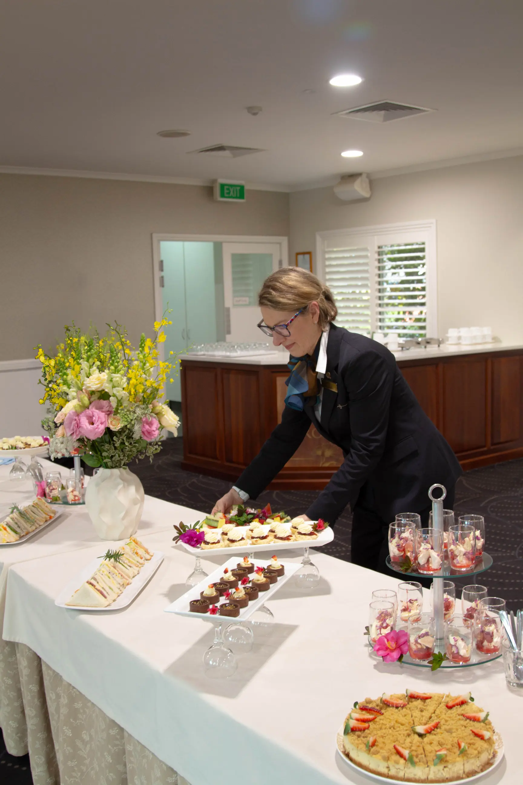 Staff member in black attire arranging food platters on a long white buffet table.