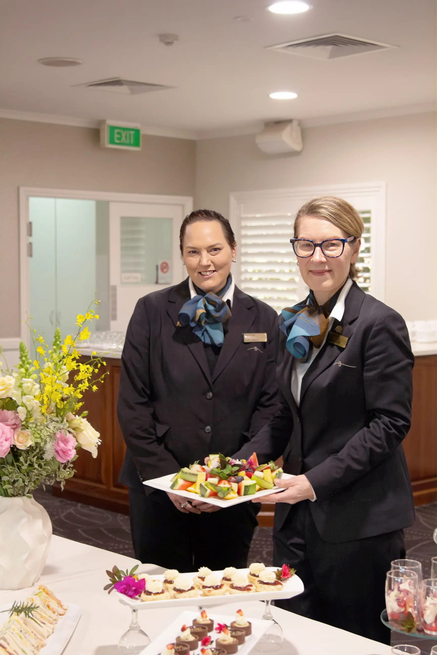 Two staff members in dark suits holding a platter of appetizers in an event room.
