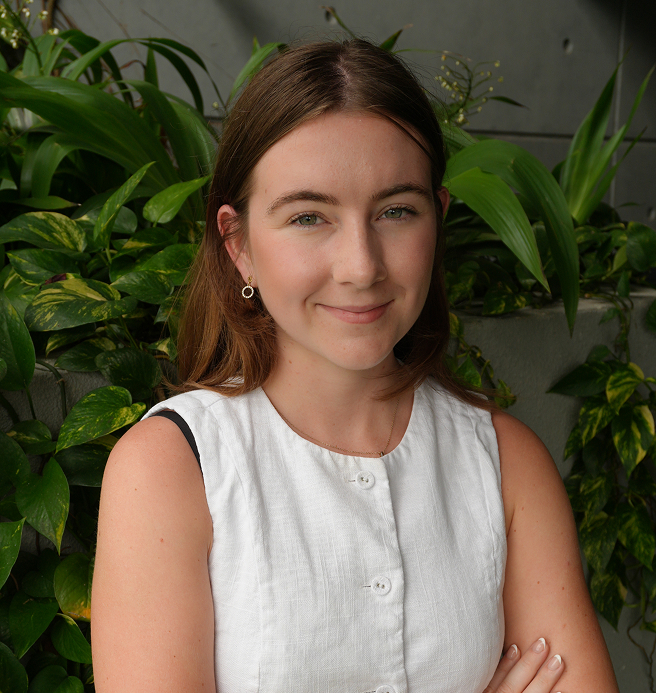Portrait of Chloe Nixon, a Webflow developer and UI/UX designer, standing in front of a leafy green backdrop. She is wearing a white sleeveless top, smiling confidently with arms gently crossed.