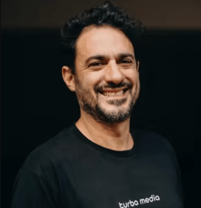 Man with dark curly hair and beard, wearing a black t-shirt with "turbo media" printed on it, smiling against a dark background.
