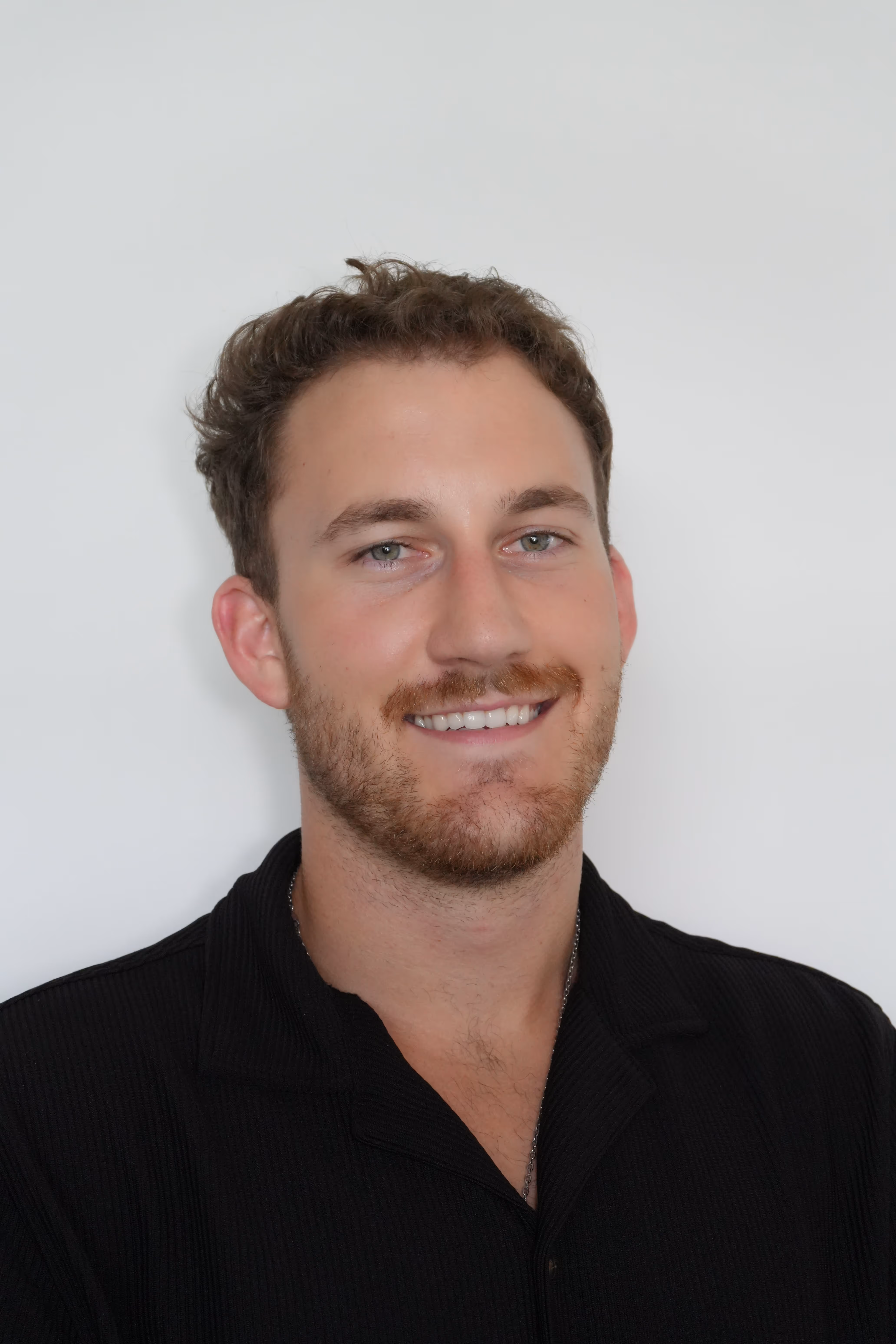 Man with short brown hair and beard wearing a black polo shirt, smiling in front of a plain white background.