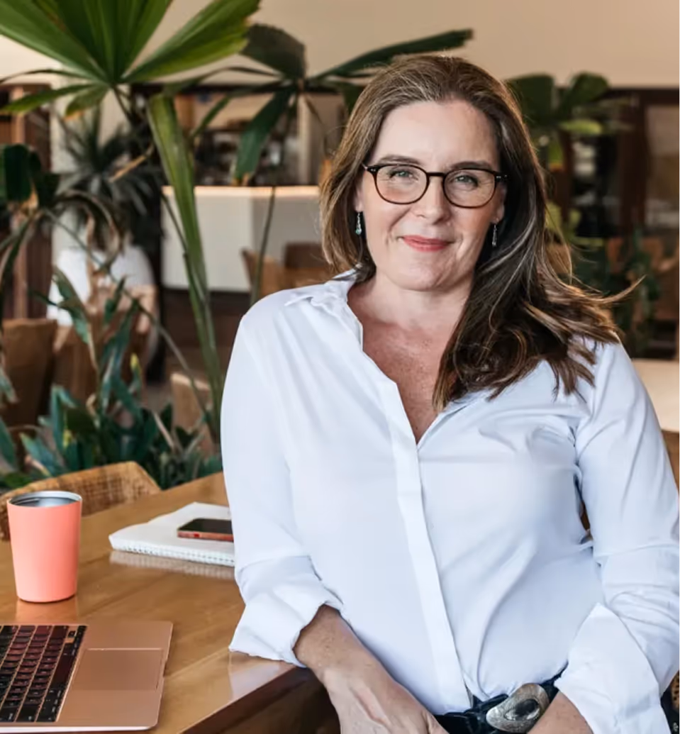 Woman in a white button-down shirt standing confidently at a wooden desk with a laptop and coffee cup, surrounded by lush indoor plants in a cozy, modern workspace.