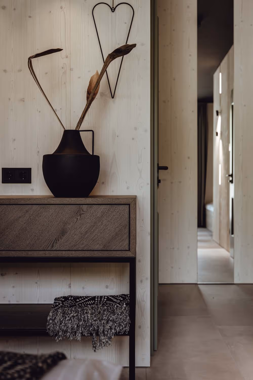 Minimalist wooden console table with a black vase holding dried plants, a black patterned throw underneath, and a heart-shaped wall decoration above.