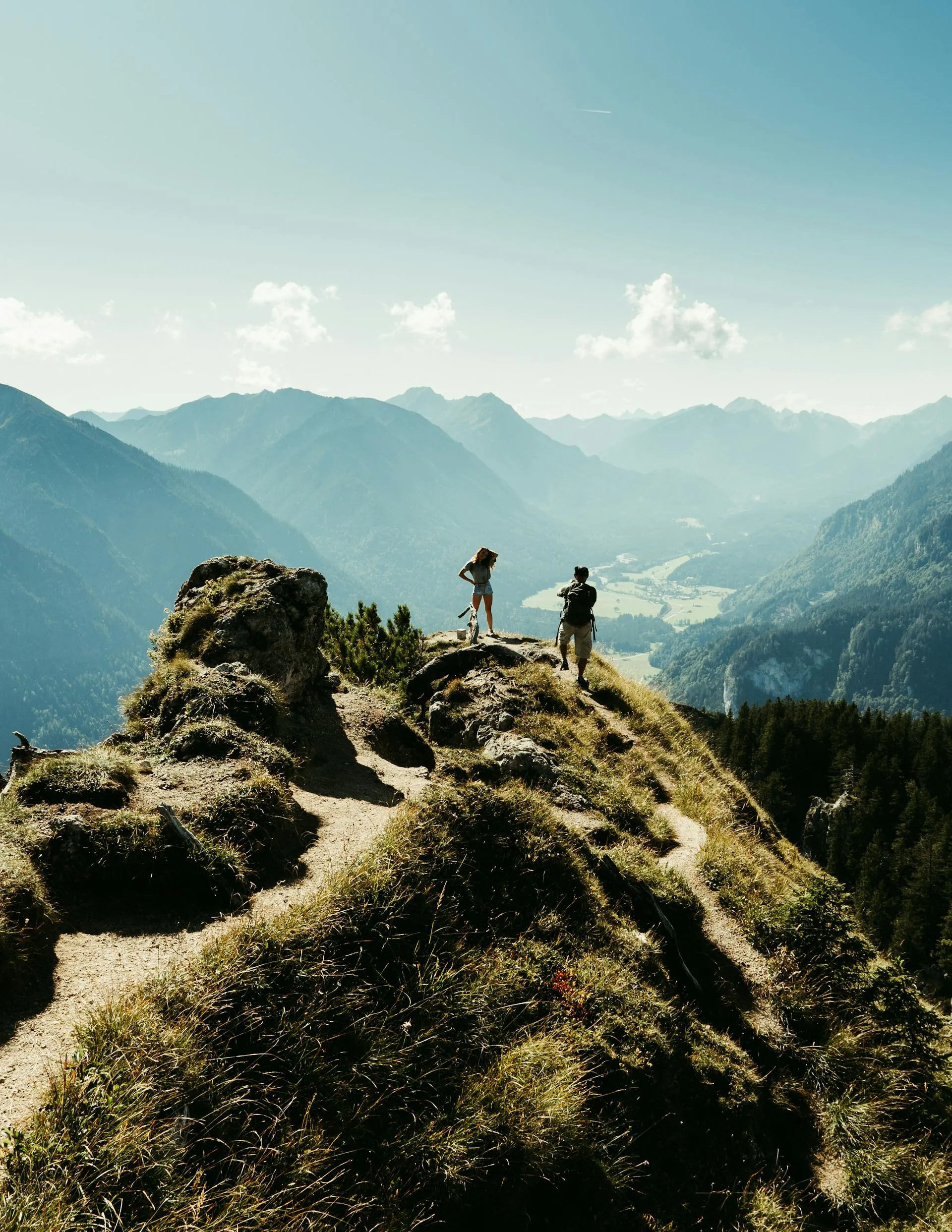 Two hikers standing on a grassy mountain ridge overlooking a valley and distant mountain ranges under a clear sky.
