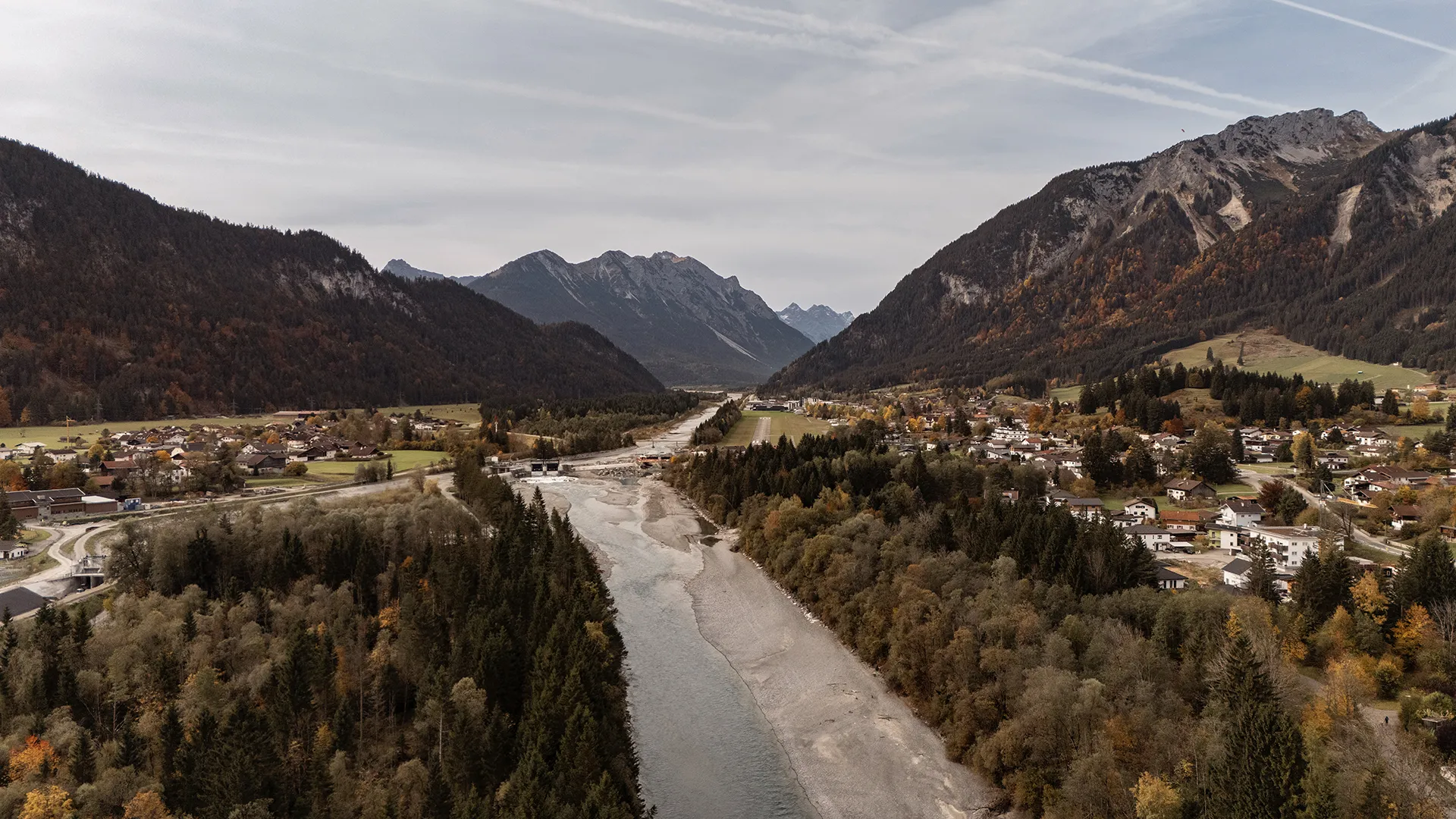 Wide river flowing through a valley with dense forest and houses on both sides, surrounded by mountains under a cloudy sky.