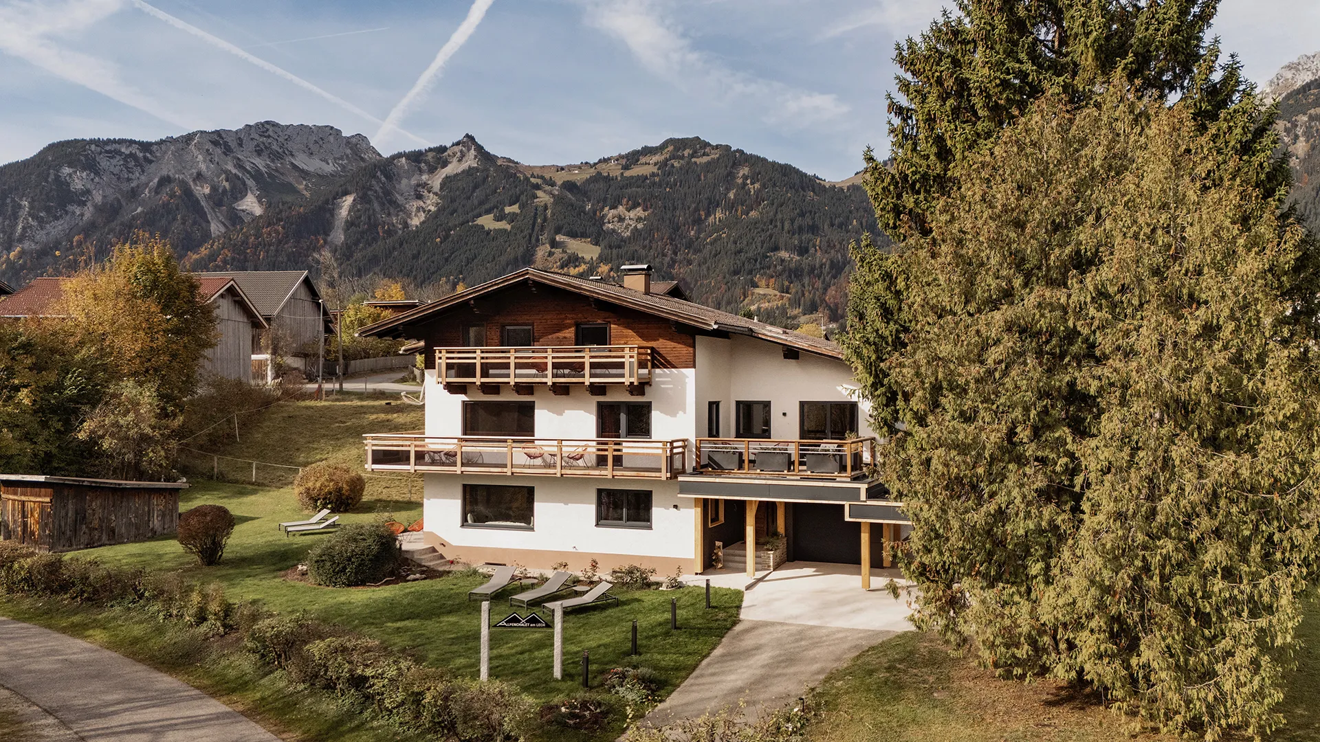 Chalet-style house with wooden balconies surrounded by lawn, large trees, and mountain backdrop under a partly cloudy sky.