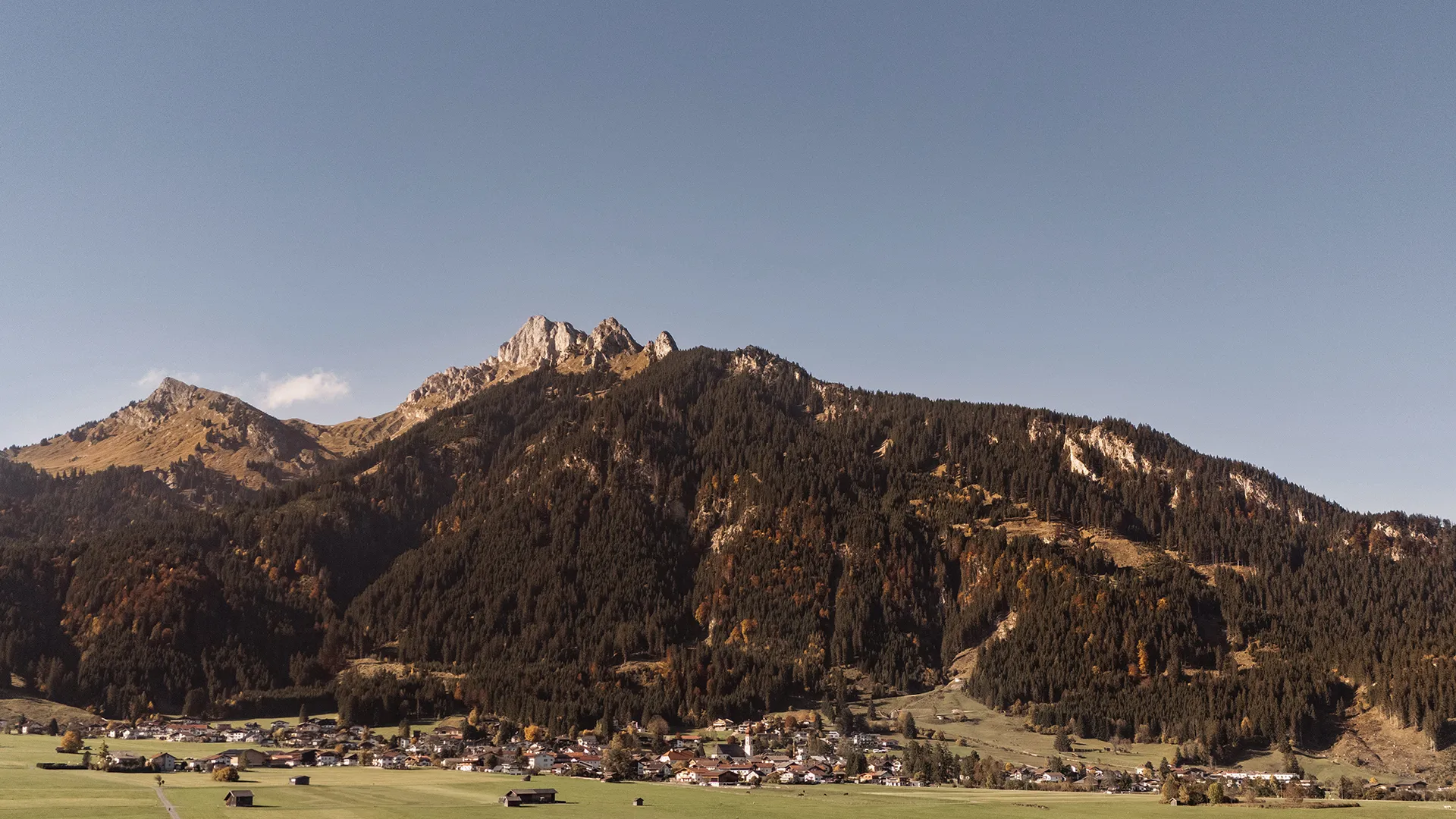Small mountain village with houses at the base of forested mountains under a clear blue sky.