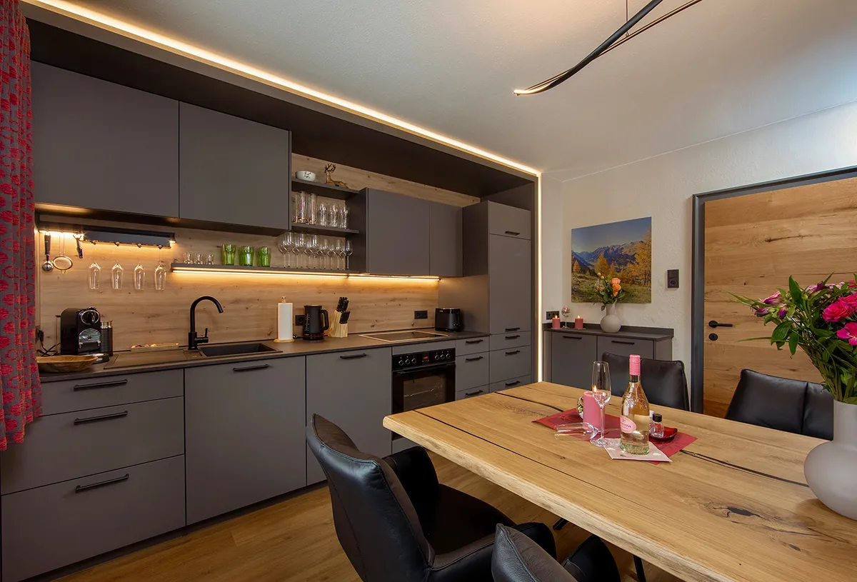 Modern kitchen with matte gray cabinets, wooden backsplash, under-cabinet lighting, a wooden dining table with black chairs, and fresh flowers in vases.