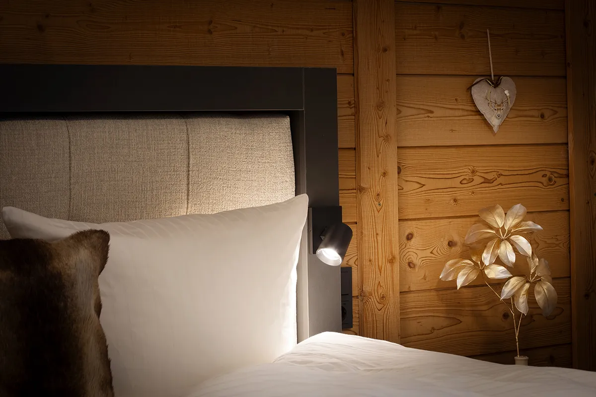 Cozy bedroom corner with beige upholstered headboard, white pillow, fur cushion, wall-mounted reading light, wooden panel wall, and gold decorative plant.