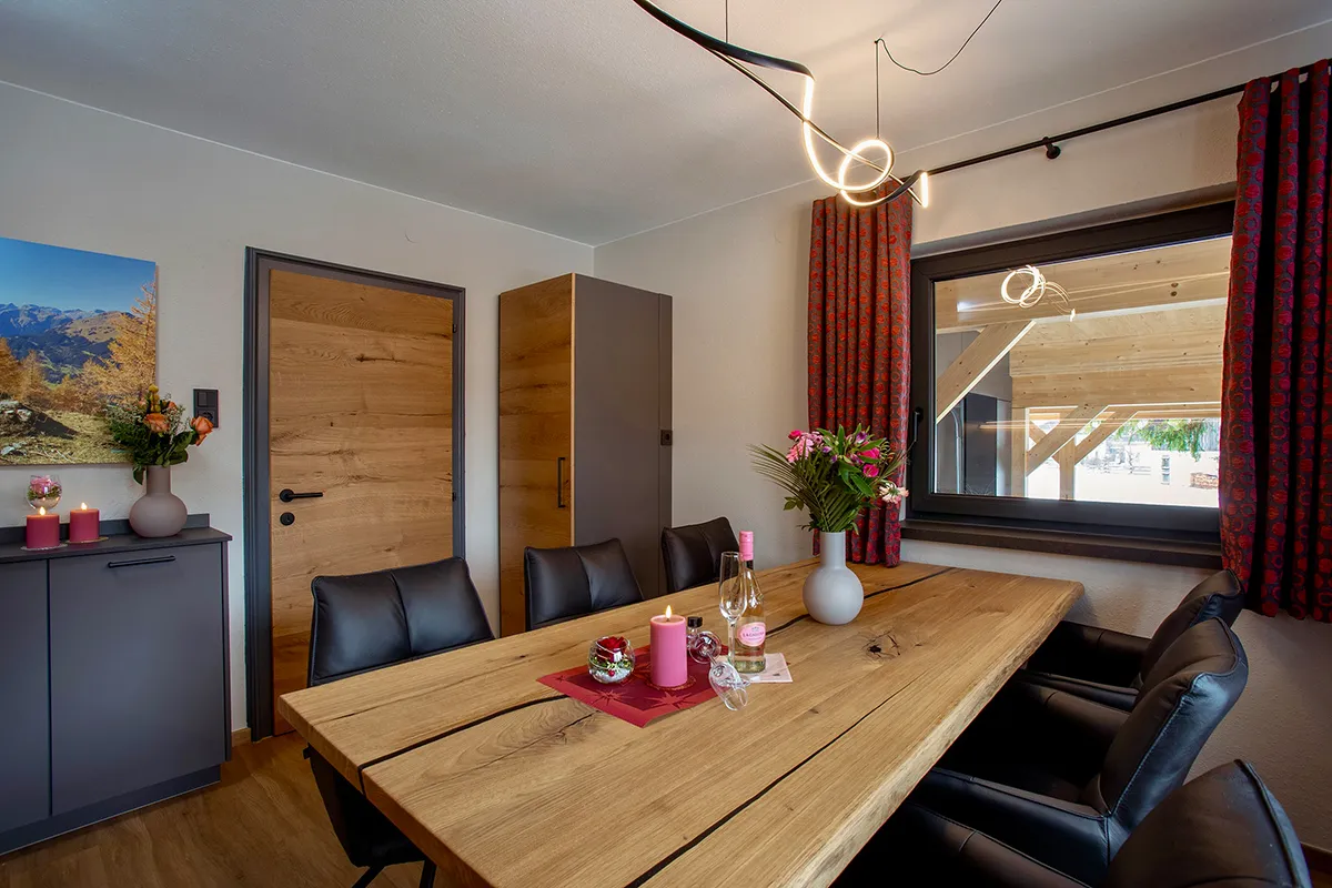 Modern dining room with a wooden table, black leather chairs, a lit pink candle, flower vases, and a large window with red curtains.
