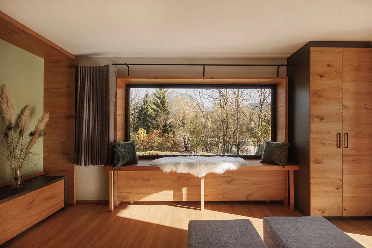 Cozy wooden interior with a large picture window showing trees and mountains, featuring a bench with fur and green pillows.