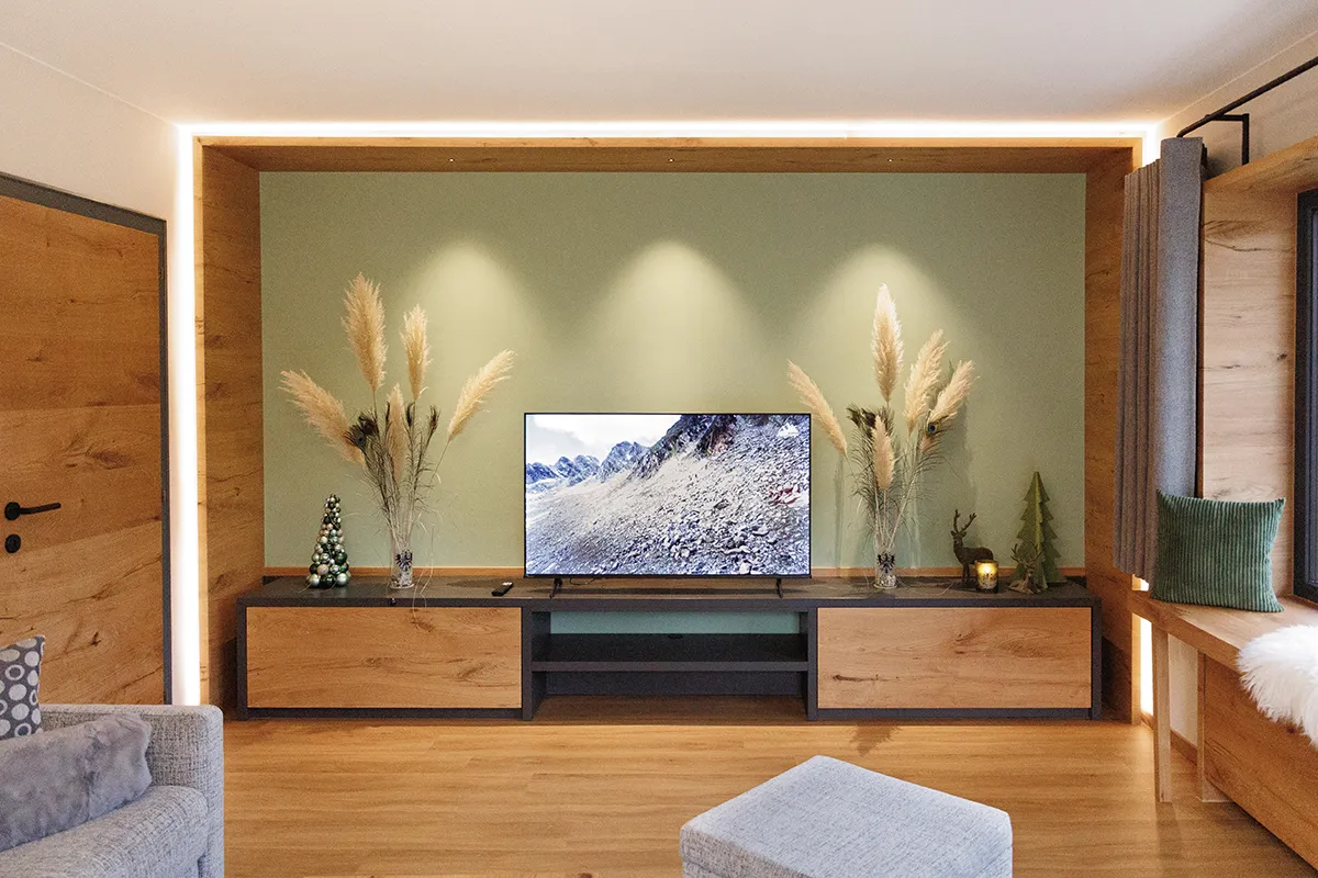 Modern living room with wooden TV stand, flat-screen TV showing mountain landscape, pampas grass in vases, small decorative Christmas trees, and cozy seating area.