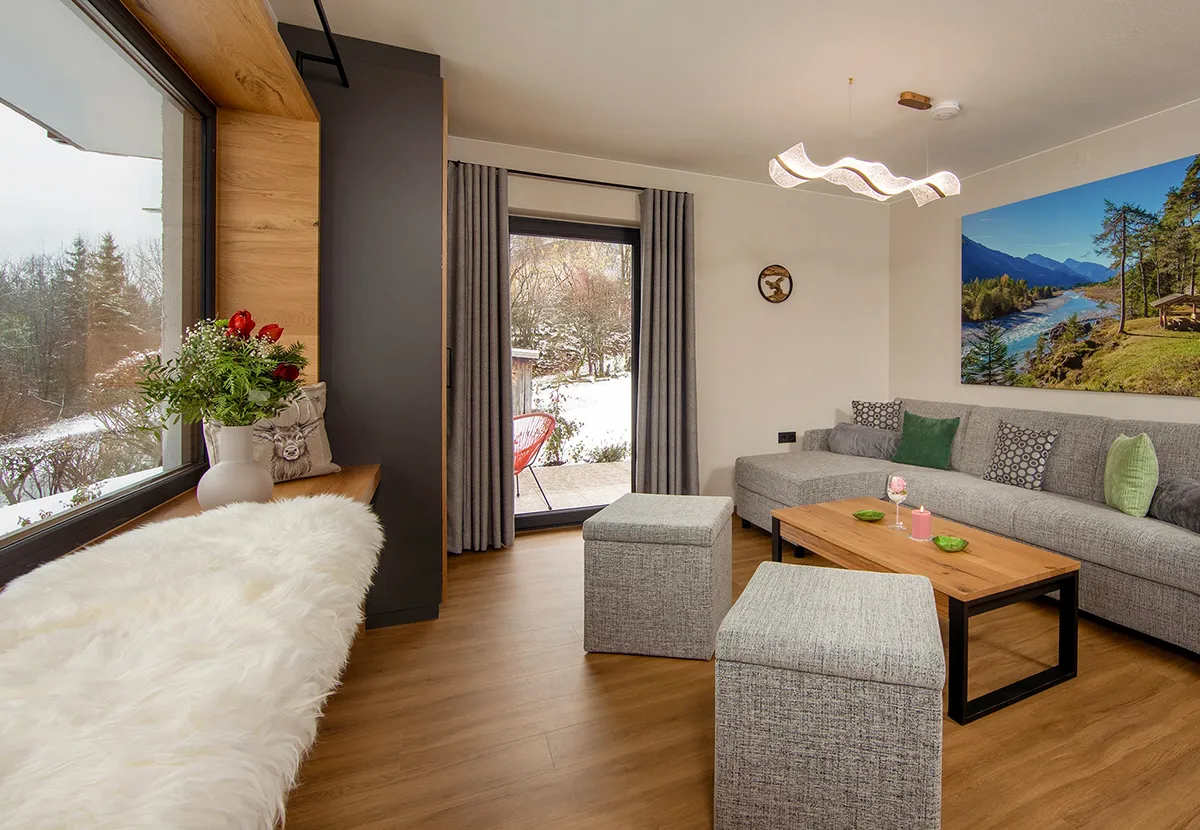 Cozy modern living room with gray sectional sofa, wooden coffee table, two gray ottomans, large window with snowy outdoor view, and a wall-mounted landscape photo.
