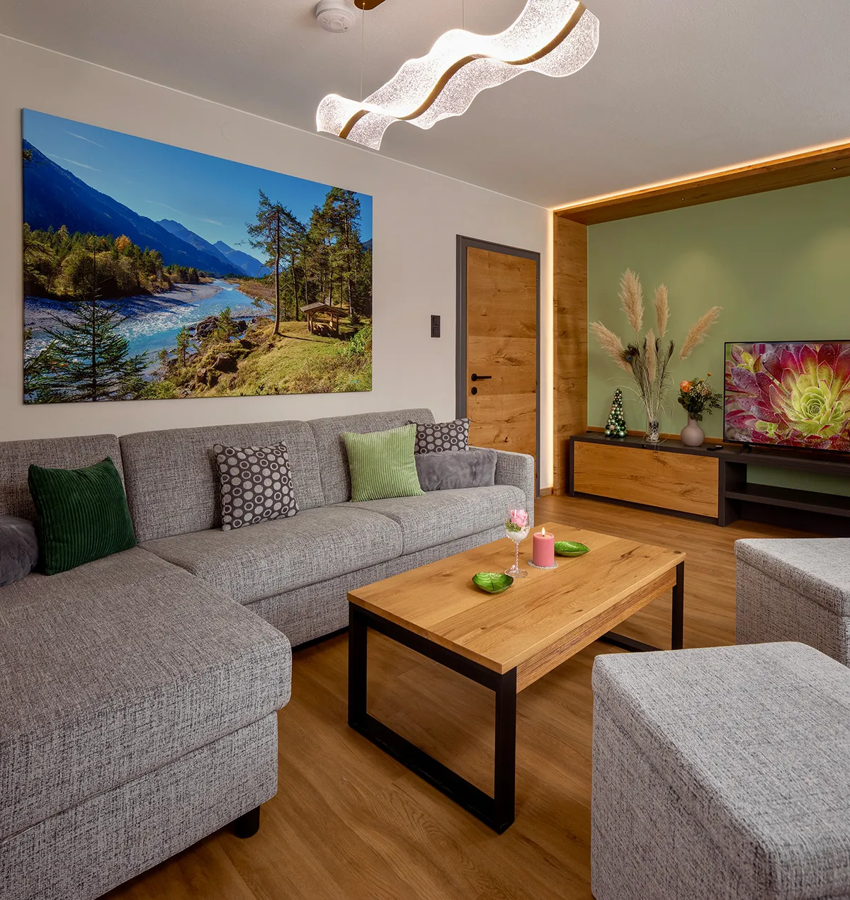 Modern living room with a gray sectional sofa, wooden coffee table with a candle and glass, wall art of a river landscape, and a TV on a wooden cabinet.