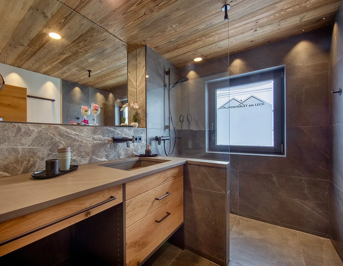 Modern bathroom with wood ceiling, gray stone walls, a large mirror, wooden vanity with sink, and a walk-in shower featuring a window with frosted glass.