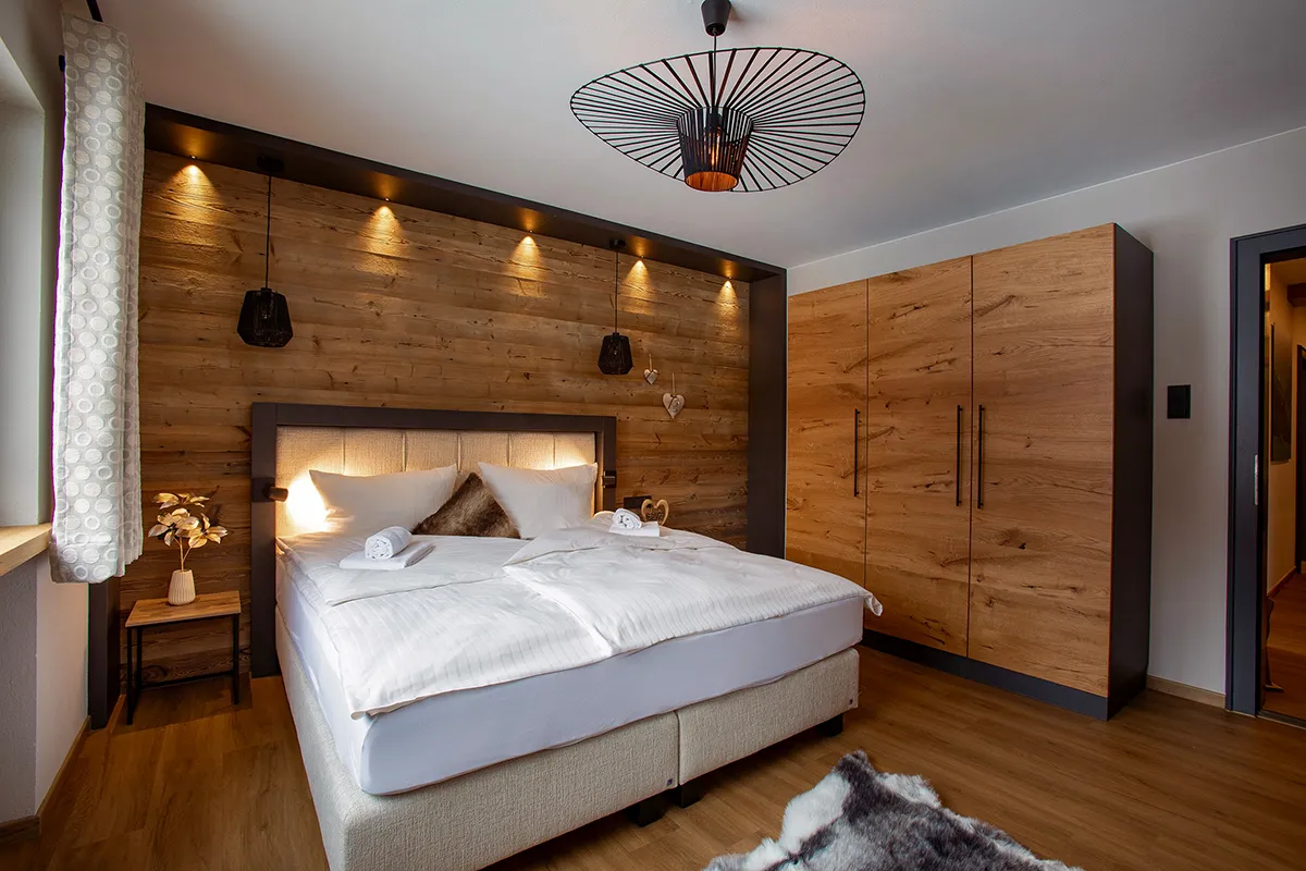 Cozy bedroom with a large bed featuring white linens, wooden wall paneling with pendant lights, wooden wardrobe, and a fur rug on wooden floor.