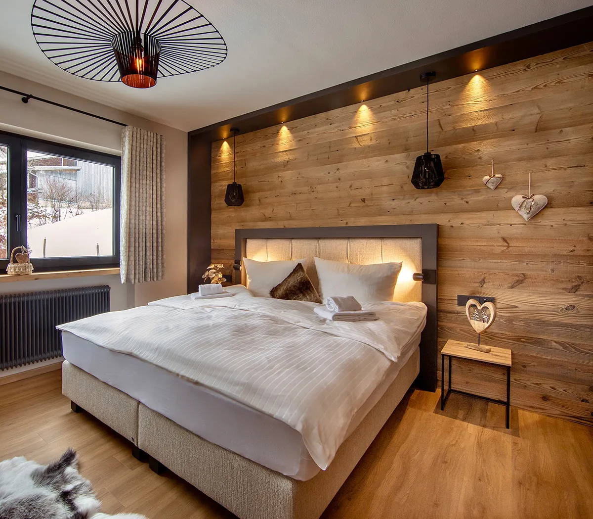 Cozy bedroom with a large bed, beige upholstered headboard with warm backlighting, wooden accent wall, two hanging pendant lights, and a small bedside table with a heart decoration.