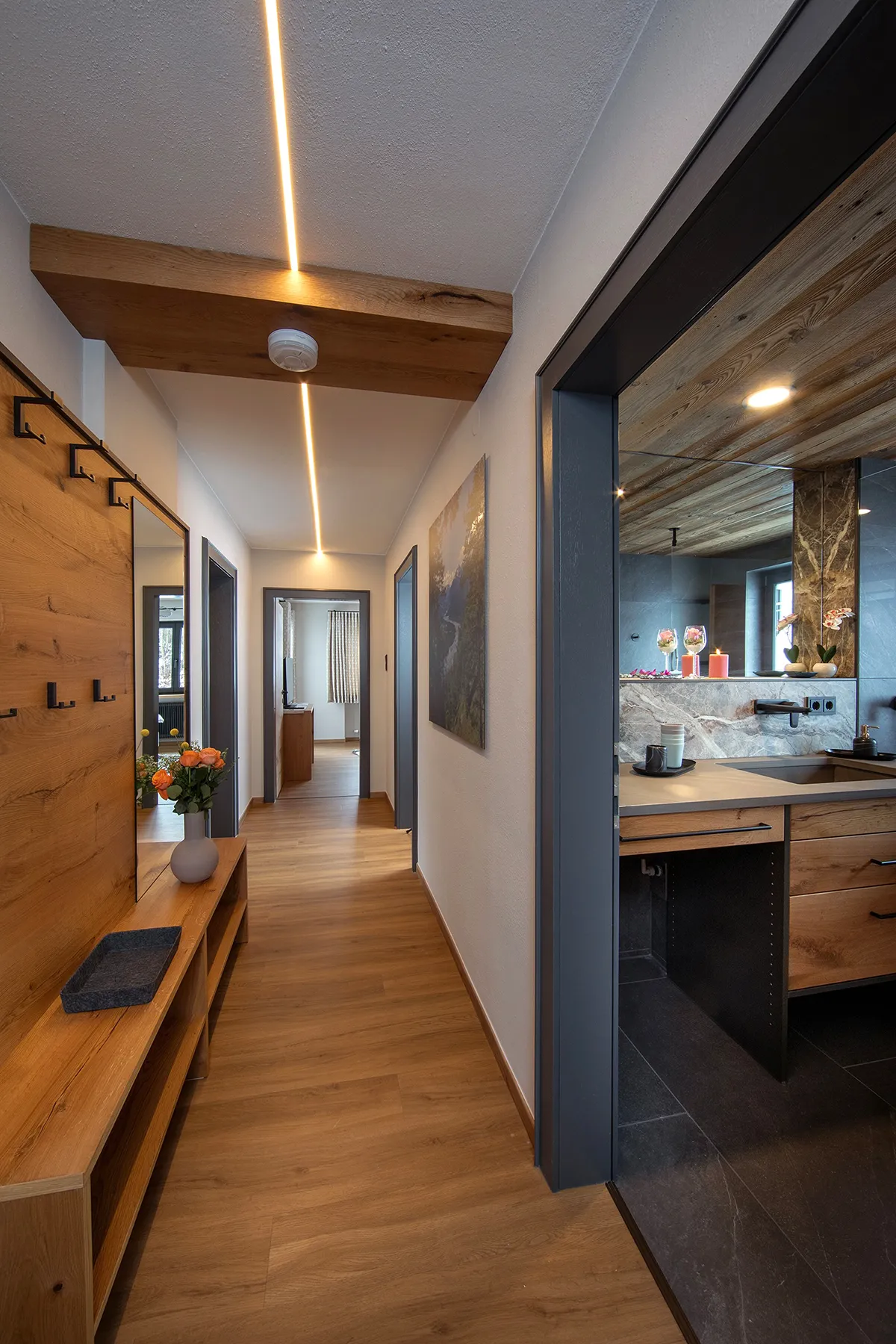 Modern hallway with wooden flooring, a wooden bench with a vase of orange flowers, coat hooks on the wall, and a view into a bathroom with wooden and stone accents.