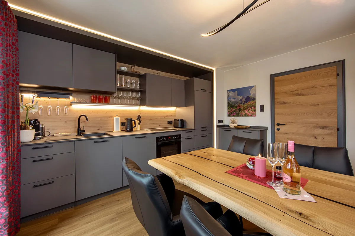 Modern kitchen with grey cabinets, wooden backsplash, and a wooden dining table set with a bottle of wine, glasses, and a lit candle.