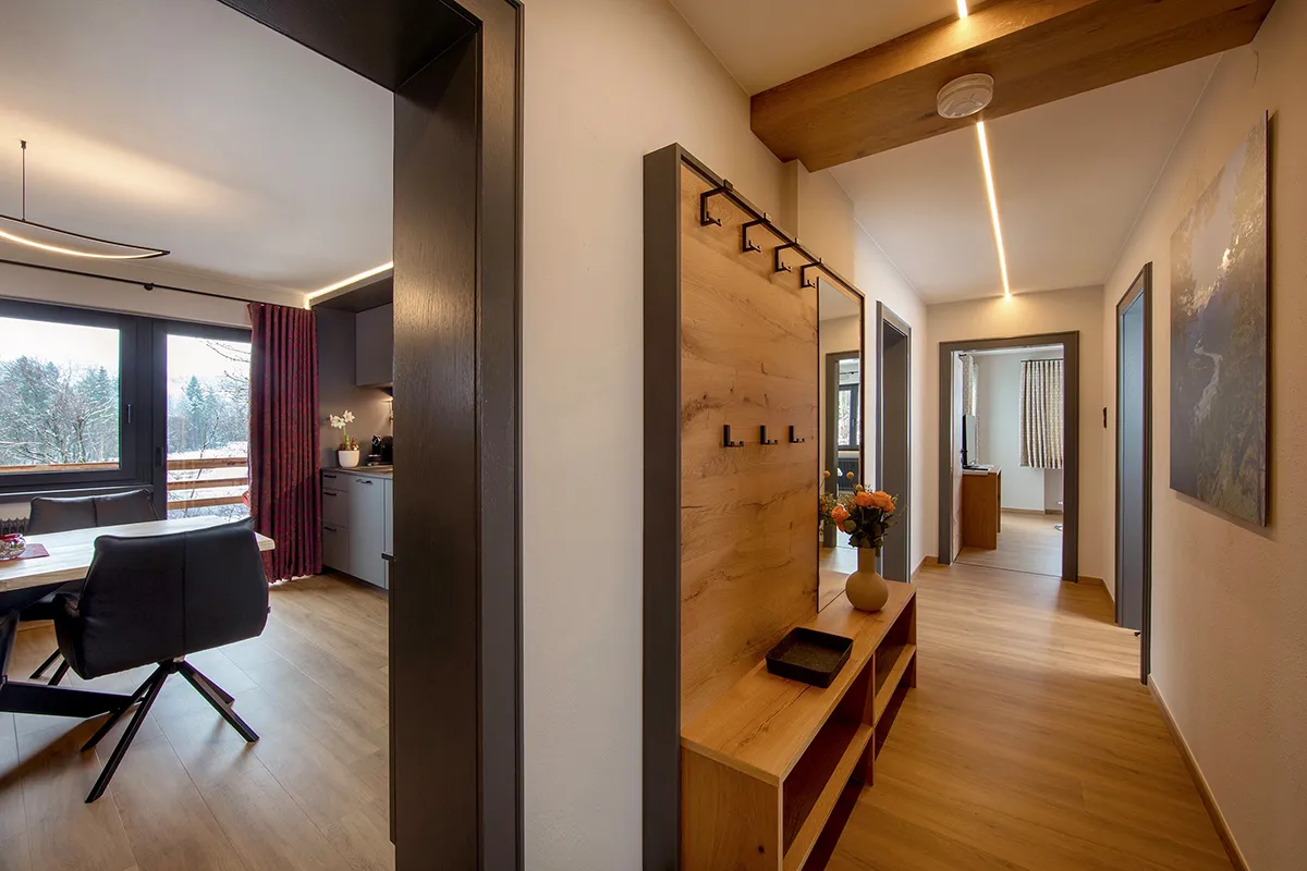 Modern hallway with wooden flooring, wall-mounted coat hooks, a mirror, and a vase of flowers on a wooden bench, leading to a room with a TV and window.