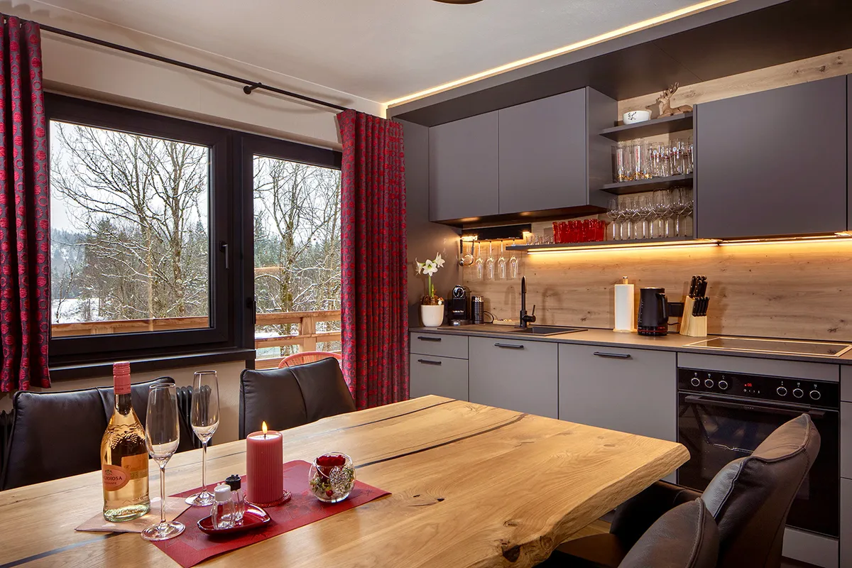 Modern kitchen with grey cabinets, wooden countertops, red patterned curtains, and a wooden dining table set with two wine glasses, a bottle, and a lit candle.
