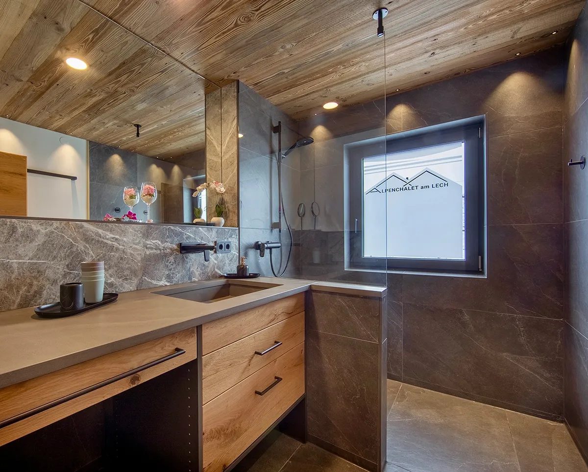 Modern bathroom with wooden ceiling, stone walls, a large mirror, a sink with countertop, and a walk-in shower with a window displaying 'Alpenchalet am Lech'.